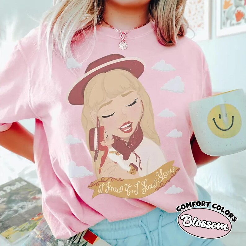 Może przedstawiać: Jasnor&oacute;żowy t-shirt z grafiką przedstawiającą blondynkę w bordowym kapeluszu trzymającą małą lalkę. Wz&oacute;r zawiera białe chmury oraz wstęgę z napisem I Knew It, I Knew You. Koszulka posiada okrągły dekolt i kr&oacute;tkie rękawy.