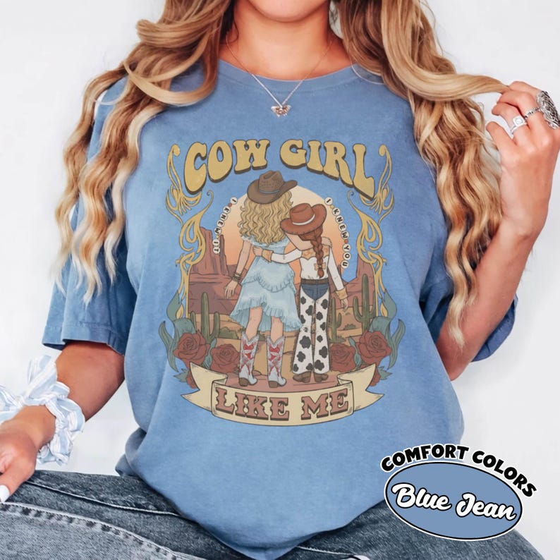 May include: A blue short-sleeve t-shirt featuring a graphic print of two figures in cowboy hats standing in a desert landscape with cacti and roses. The text reads COW GIRL LIKE ME. The shirt has a vintage-style wash and a crew neckline.