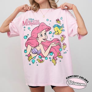 Puede incluir: Una camiseta rosa claro con un estampado gr&aacute;fico de Ariel de La Sirenita de Disney, junto a Flounder y varios caballitos de mar rodeados de burbujas turquesas. El texto Disney The Little Mermaid est&aacute; impreso en la esquina superior izquierda.