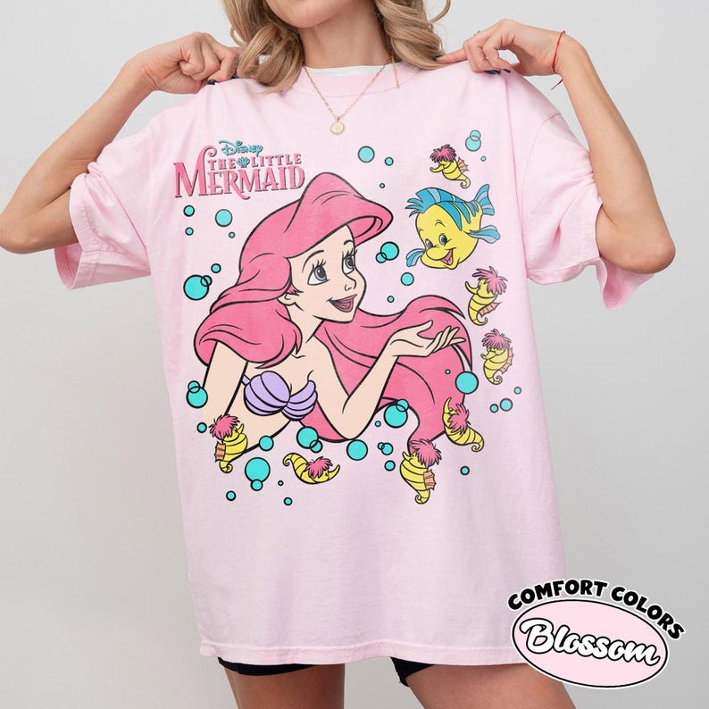 Puede incluir: Una camiseta rosa claro con un estampado gr&aacute;fico de Ariel de La Sirenita de Disney, junto a Flounder y varios caballitos de mar rodeados de burbujas turquesas. El texto Disney The Little Mermaid est&aacute; impreso en la esquina superior izquierda.