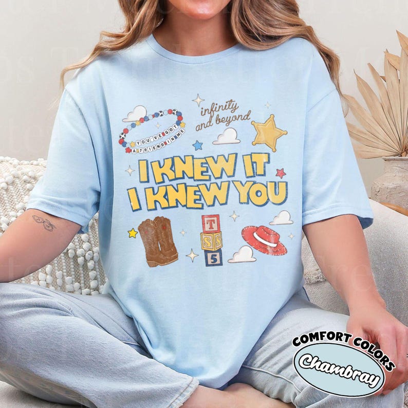 May include: A light blue t-shirt featuring a graphic print with the text "I KNEW IT I KNEW YOU" and "infinity and beyond". The design includes a beaded bracelet, a yellow star, brown cowboy boots, alphabet blocks, and a red cowboy hat.