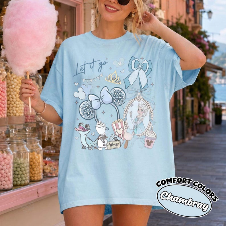 May include: A light blue t-shirt featuring a graphic print with the text Let it go, a character in a mirror, a snowman, a lizard, mouse ears, a bow, and a popcorn bucket. The design includes various snowflake and star accents in shades of blue and white.