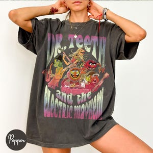 Könnte beinhalten: Dunkelgraues T-Shirt mit einem farbenfrohen Grafikdesign von Dr. Teeth und der Band Electric Mayhem. Das Design zeigt verschiedene Muppet-Charaktere, die Instrumente spielen. Der Text "Dr. Teeth and the Electric Mayhem" ist in einem Farbverlauf gedruckt.