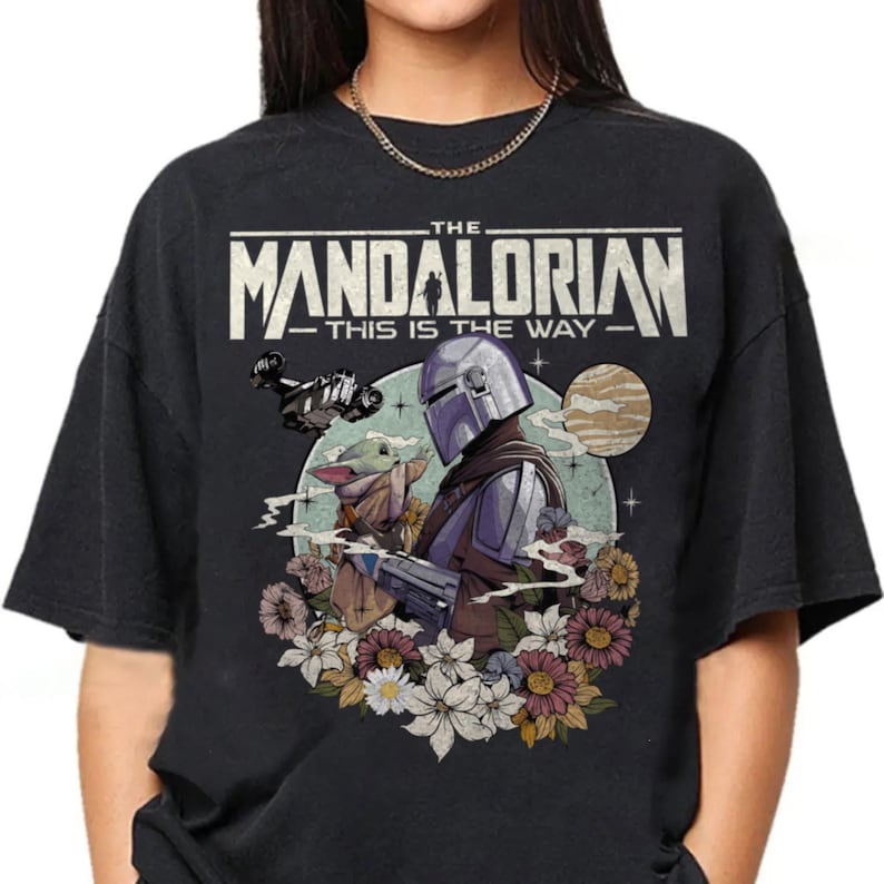 Op de afbeelding: Een zwart t-shirt met een grafische print van The Mandalorian en Grogu. De tekst luidt THE MANDALORIAN THIS IS THE WAY. Het ontwerp bevat een ruimteschip, een planeet en een groep kleurrijke bloemen onderaan in een cirkelvormig kader.