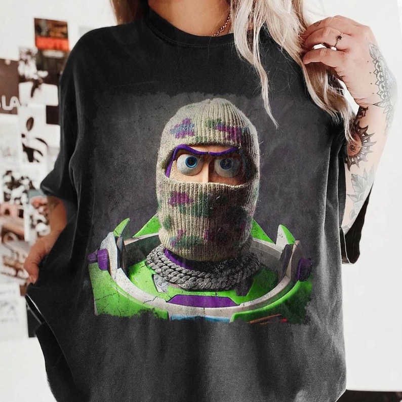 K&ouml;nnte beinhalten: Ein schwarzes T-Shirt mit einem Grafikdruck einer Figur, die eine beigefarbene Stricksturmhaube mit violetten und gr&uuml;nen Tarnmustern tr&auml;gt. Die Figur hat blaue Augen und tr&auml;gt eine silberne Kette &uuml;ber einem gr&uuml;n-violetten Raumanzugkragen.