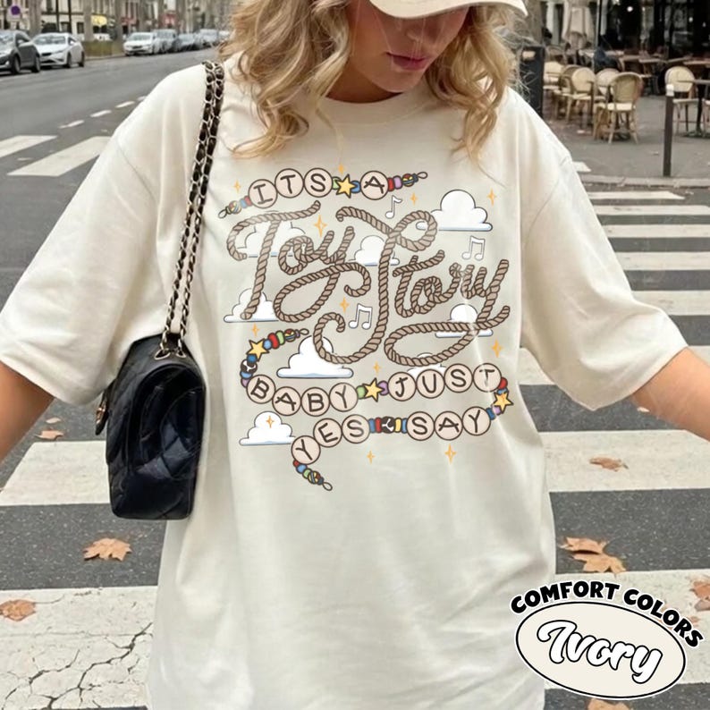 May include: An ivory t-shirt featuring a graphic print with the text "IT'S A TOY STORY BABY JUST YES SAY" written in a rope-style font. The design includes colorful beads, small stars, musical notes, and white clouds on a light background.