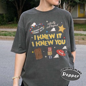 Pode incluir: Uma t-shirt cinzento-carv&atilde;o de manga curta com uma impress&atilde;o gr&aacute;fica com o texto "I KNEW IT I KNEW YOU" e "infinity and beyond". O design inclui uma pulseira de contas, uma estrela amarela, uma bota castanha, blocos e um chap&eacute;u vermelho.
