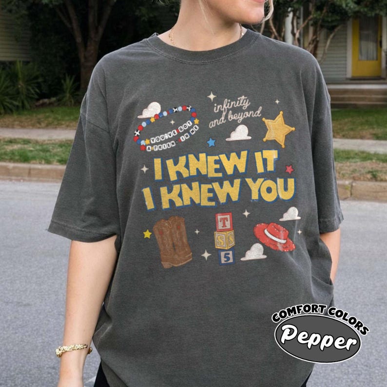 Pode incluir: Uma t-shirt cinzento-carv&atilde;o de manga curta com uma impress&atilde;o gr&aacute;fica com o texto "I KNEW IT I KNEW YOU" e "infinity and beyond". O design inclui uma pulseira de contas, uma estrela amarela, uma bota castanha, blocos e um chap&eacute;u vermelho.