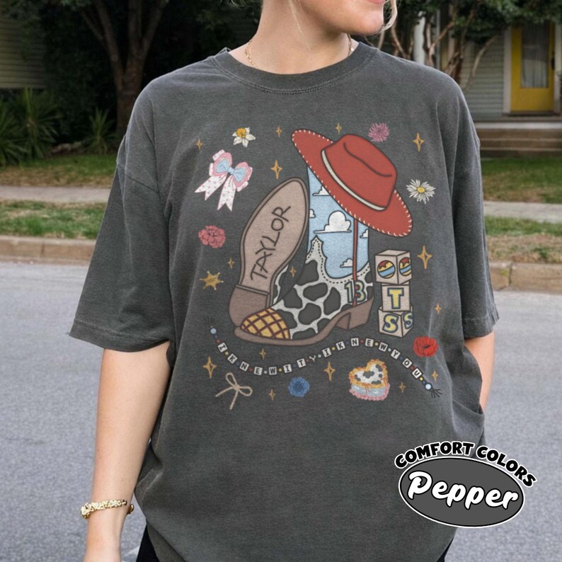 Op de afbeelding: Een antracietgrijs t-shirt met een grafische print van een cowboylaars met koeienprint, een rode hoed en blokken met de letters T S. Het ontwerp bevat de tekst TAYLOR, het getal 13 en een kralenketting met de tekst I KNEW IT I KNEW YOU.