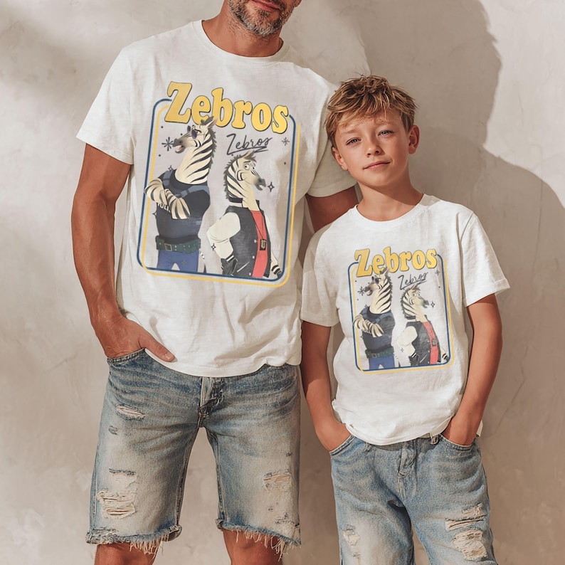 May include: Two white t-shirts featuring the word "Zebros" in yellow and a cartoon zebra design. A man and a child wear the shirts, paired with denim shorts. The background is a plain, light-coloured wall.