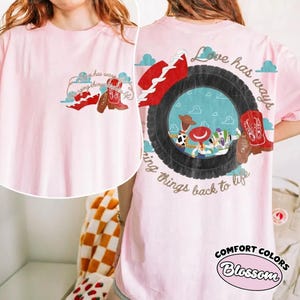May include: A light pink t-shirt featuring a small graphic on the front left chest and a large graphic on the back. Both graphics show a black tire, a red cowboy boot, and the text Love has ways of bringing things back to life.