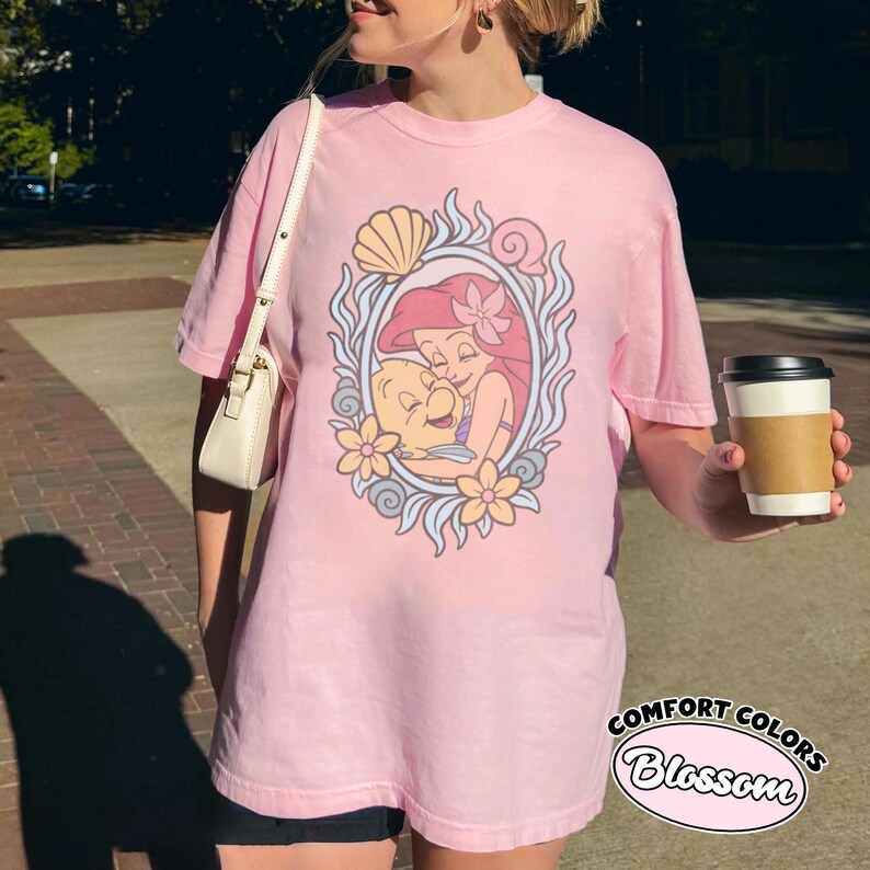 May include: A light pink short-sleeve t-shirt featuring a graphic of a cartoon mermaid and a small fish enclosed in an oval frame decorated with shells and flowers. The shirt has a crew neckline and a relaxed fit, with a white shoulder bag strap visible.