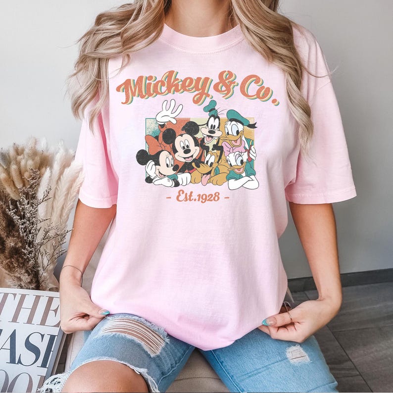 以下が含まれることがあります： ミッキーマウスと仲間たちのレトロなグラフィックが特徴のライトピンクのTシャツ。「Mickey & Co.」と「Est. 1928」の文字が入っています。半袖でゆったりとしたフィット感です。