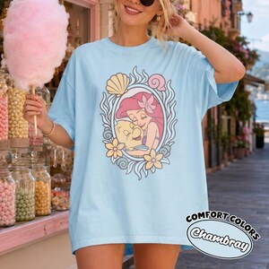 May include: A light blue t-shirt featuring a graphic print of a red-haired mermaid character hugging a yellow fish. The illustration is framed by an oval border decorated with seashells and flowers. The shirt has a loose, casual fit.