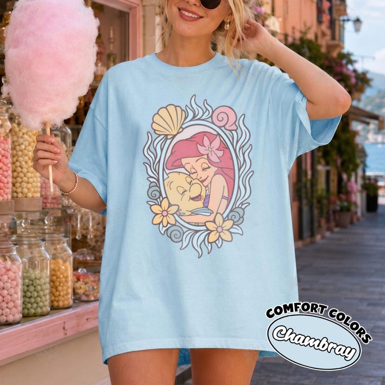 May include: A light blue t-shirt featuring a graphic print of a red-haired mermaid character hugging a yellow fish. The illustration is framed by an oval border decorated with seashells and flowers. The shirt has a loose, casual fit.