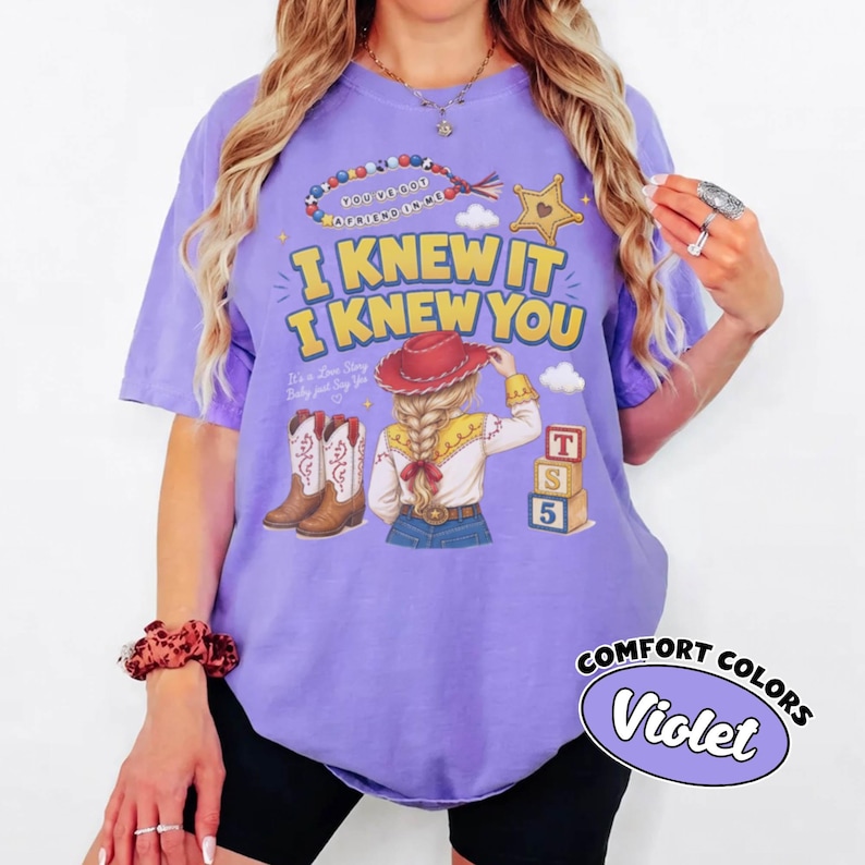 May include: A violet short-sleeve t-shirt featuring a graphic print of a cowgirl with a red hat, cowboy boots, and alphabet blocks. The text reads I KNEW IT I KNEW YOU, along with a beaded bracelet graphic and a yellow star. The shirt is labelled Comfort Colors.