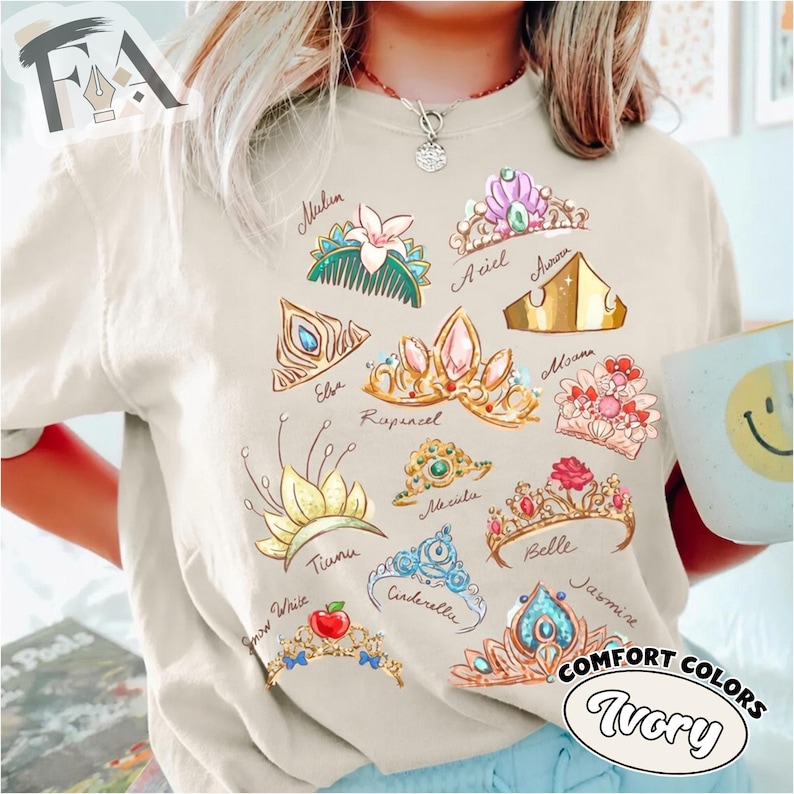 May include: Ivory-colored t-shirt featuring watercolor illustrations of various princess crowns, each labeled with a name like Ariel, Belle, and Cinderella. The shirt also has the words "Comfort Colors" and "Ivory" printed on it.