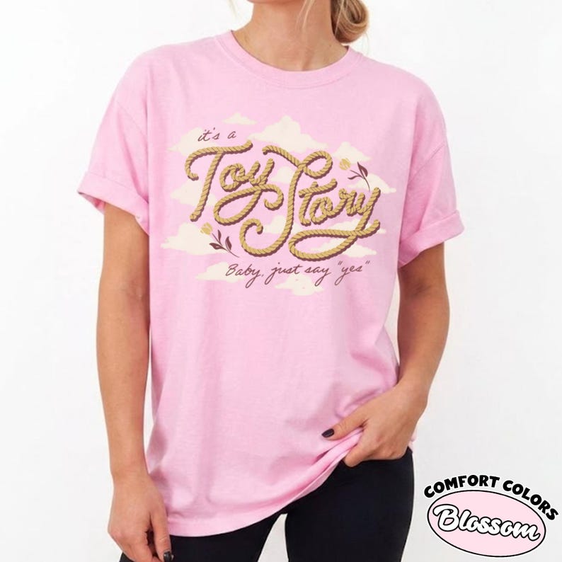 May include: A light pink short-sleeve t-shirt featuring a graphic of tan rope styled into the text "Toy Story" against a background of pale yellow clouds. Below the rope text, the phrase "it's a" and "Baby, just say "yes"" are printed.