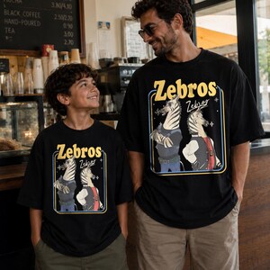 May include: Two black t-shirts featuring the text "Zebros" in yellow and a cartoon zebra design. The design shows two zebras wearing clothes. The shirts are worn by a man and a boy.