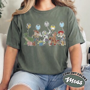 May include: Moss green Comfort Colors t-shirt with a Toy Story character graphic holding balloons. Characters include Rex, Woody, Buzz Lightyear, and Jessie. The shirt has a relaxed fit, and the words "Comfort Colors" and "Moss" are printed on the bottom right.