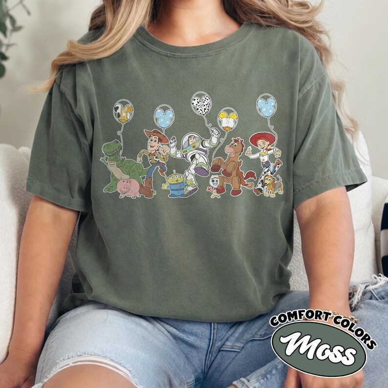 May include: Moss green Comfort Colors t-shirt with a Toy Story character graphic holding balloons. Characters include Rex, Woody, Buzz Lightyear, and Jessie. The shirt has a relaxed fit, and the words "Comfort Colors" and "Moss" are printed on the bottom right.