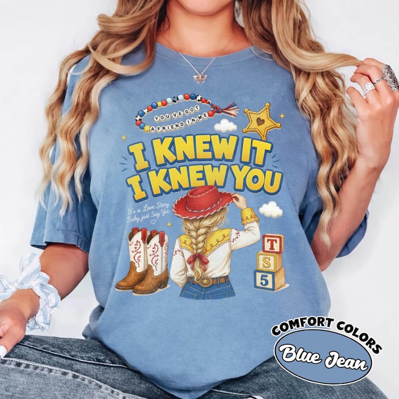 May include: A blue denim-coloured t-shirt featuring a graphic print of a person with a blonde braid wearing a red cowboy hat, brown cowboy boots, and a yellow shirt. The text reads "I KNEW IT I KNEW YOU", "YOU'VE GOT A FRIEND IN ME", and "It's a Love Story Baby just Say Yes".