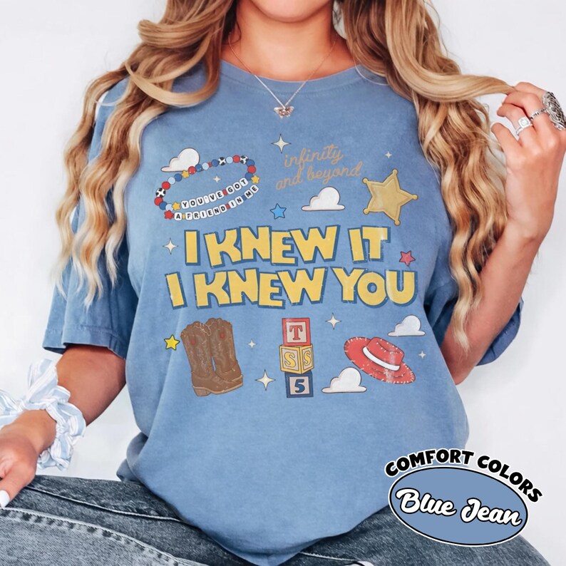 May include: A blue jean coloured t-shirt featuring yellow text that reads I KNEW IT I KNEW YOU. The graphic includes a beaded bracelet with the text YOU'VE GOT A FRIEND IN ME, a gold star, brown cowboy boots, alphabet blocks, and a red hat.