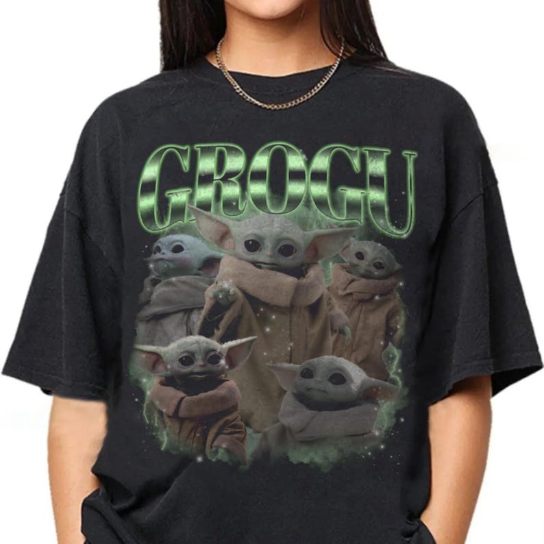 May include: A black short-sleeved t-shirt featuring a graphic print of five small green creatures with large ears and tan robes. The text GROGU is printed in a large, stylized green font above the characters, surrounded by a faint green mist.