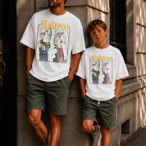May include: Two people wearing matching white t-shirts with a cartoon zebra design and the text "Zebros". They are also wearing olive green shorts. The image is taken outdoors.