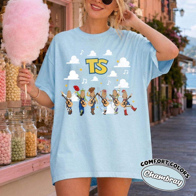 Może przedstawiać: Jasnoniebieski t-shirt z grafiką przedstawiającą sześć postaci z gitarami pod białymi chmurami oraz ż&oacute;łtym napisem TS. Wz&oacute;r zawiera nuty i ż&oacute;łte gwiazdki. Koszulka ma kr&oacute;tkie rękawy i okrągły dekolt.