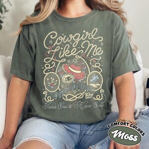 以下が含まれることがあります： モスグリーンの半袖Tシャツで、Cowgirl Like MeおよびTakes One to Know Oneというテキストのグラフィックプリントが施されています。デザインには、赤いカウボーイハット、蹄鉄、ロープの縁取り、音符、小さな星のアクセントが含まれています。