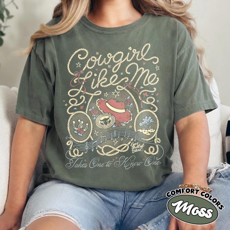 以下が含まれることがあります： モスグリーンの半袖Tシャツで、Cowgirl Like MeおよびTakes One to Know Oneというテキストのグラフィックプリントが施されています。デザインには、赤いカウボーイハット、蹄鉄、ロープの縁取り、音符、小さな星のアクセントが含まれています。