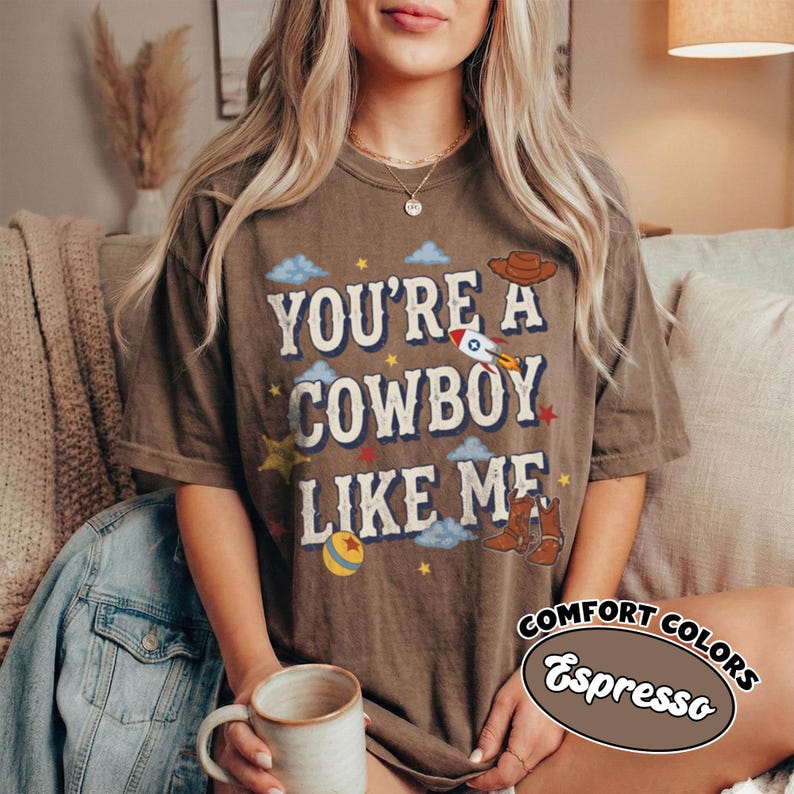 May include: A brown short-sleeve t-shirt featuring the text "YOU'RE A COWBOY LIKE ME" in a white western-style font. The design includes illustrations of a rocket, a cowboy hat, cowboy boots, clouds, stars, and a ball in blue, red, and yellow.