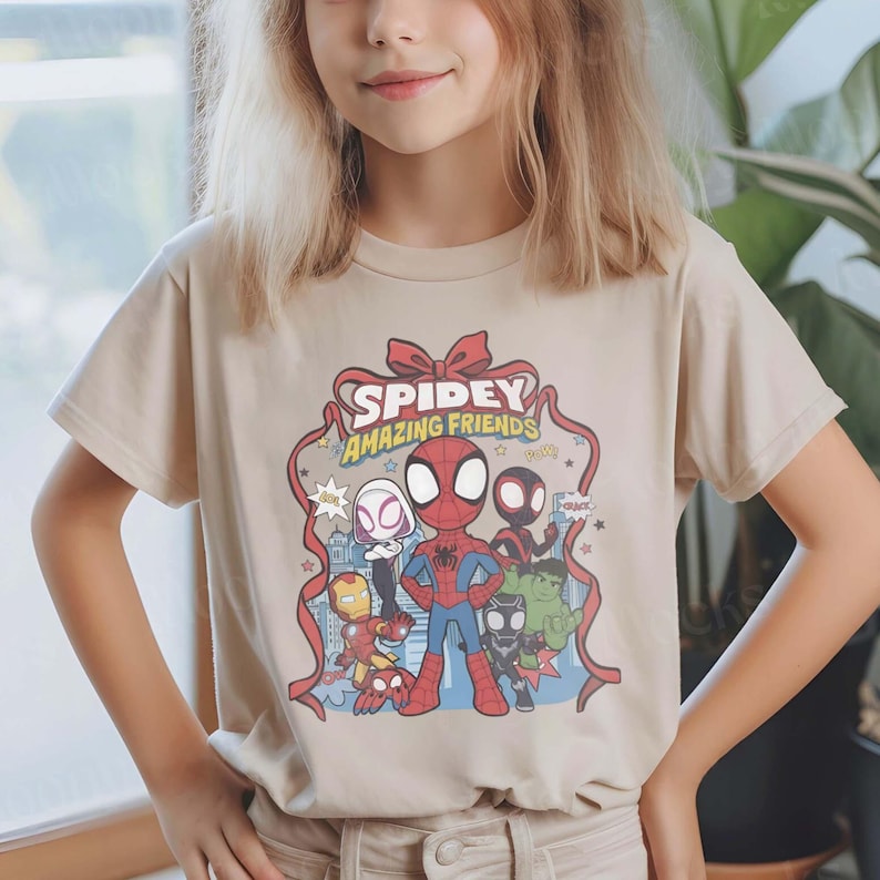 May include: Beige t-shirt featuring a colorful graphic of Spider-Man and other characters with the text "Spidey Amazing Friends". The design includes Spider-Man, Spider-Gwen, Iron Man, and the Hulk. The shirt has short sleeves.