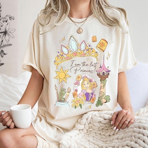 May include: Cream-colored t-shirt featuring a colorful graphic design with the text "I am the lost Princess!". The design includes a crown, sun, tower, and a princess. The shirt has short sleeves and a relaxed fit.