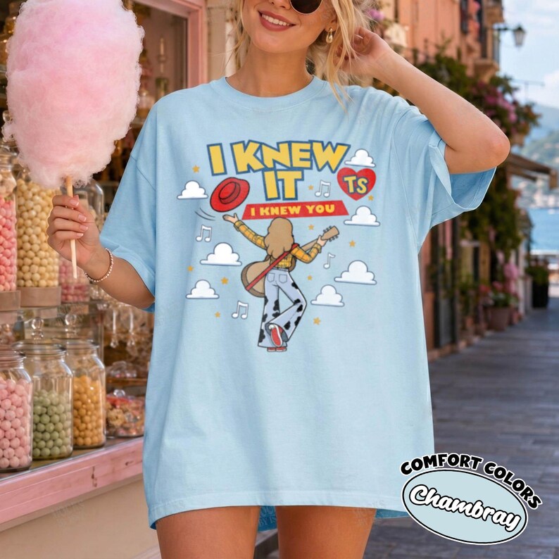 May include: A light blue t-shirt featuring a graphic of a person with long hair playing an acoustic guitar, wearing cow-print trousers and a red hat. The text reads I KNEW IT and I KNEW YOU, surrounded by clouds, musical notes, and a red heart.