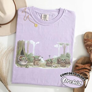 Puede incluir: Una camiseta de color lila claro con un dibujo de un personaje de dibujos animados en un bosque. La camiseta tiene las palabras "COMFORT COLORS" y "Orchid" impresas. Una cadena de oro, un sombrero y sandalias tambi&eacute;n est&aacute;n en la imagen.