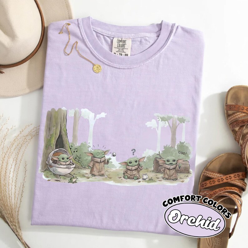 Puede incluir: Una camiseta de color lila claro con un dibujo de un personaje de dibujos animados en un bosque. La camiseta tiene las palabras "COMFORT COLORS" y "Orchid" impresas. Una cadena de oro, un sombrero y sandalias tambi&eacute;n est&aacute;n en la imagen.