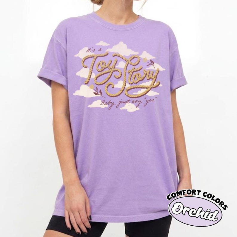 May include: A light purple short-sleeve t-shirt featuring a graphic print of white clouds and the text "It's a Toy Story" in a tan rope-style font, with the additional text "Baby, just say 'yes'" printed below in a smaller brown script font.