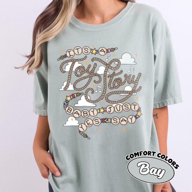 May include: A sage green t-shirt featuring a graphic design with the text "ITS A Toy Story BABY JUST YES SAY" written in a rope-style font. The design includes colorful beads, small yellow stars, musical notes, and white clouds on a light background.