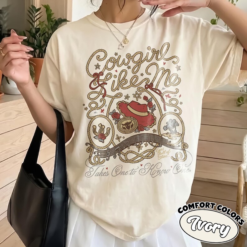 以下が含まれることがあります： アイボリーのTシャツで、ロープのデザイン、赤いカウボーイハット、バラ、音符のグラフィックプリントが施されています。テキストには「Cowgirl like Me」と「Takes One to Know One」と書かれています。黒いハンドバッグと白いスカートを合わせています。