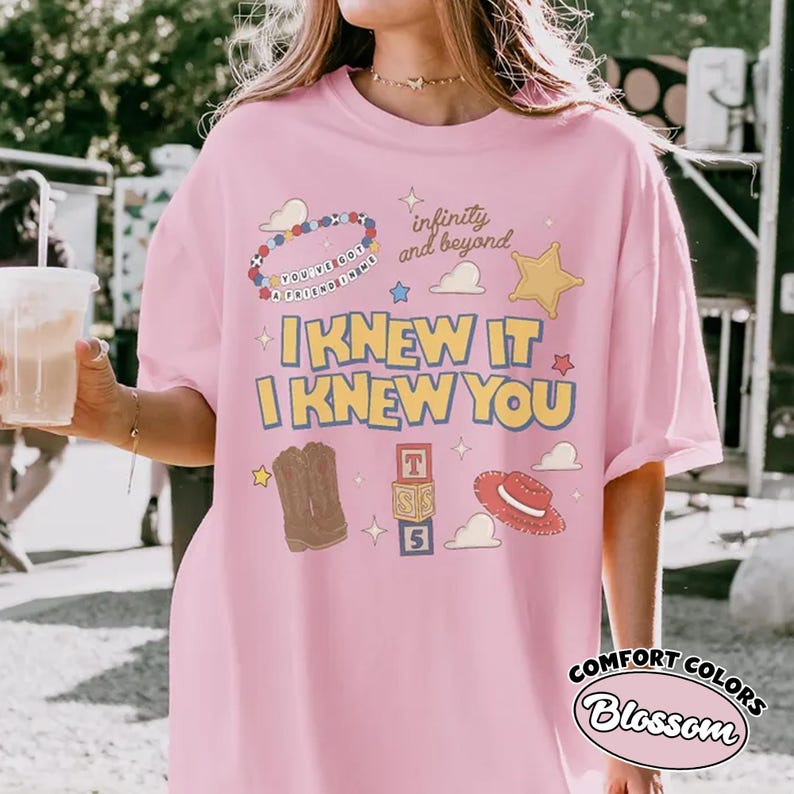 May include: A light pink t-shirt featuring a graphic print with the text "I KNEW IT I KNEW YOU", "infinity and beyond", and "YOU'VE GOT A FRIEND IN ME". The design includes illustrations of a beaded bracelet, cowboy boots, blocks, and a hat.
