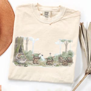 Puede incluir: Camiseta de color crema con una ilustraci&oacute;n de dibujos animados de Grogu de The Mandalorian. El gr&aacute;fico muestra a Grogu en varias poses, con un fondo de bosque. La camiseta tiene cuello redondo y mangas cortas. La etiqueta de la marca "Comfort Colors" es visible.