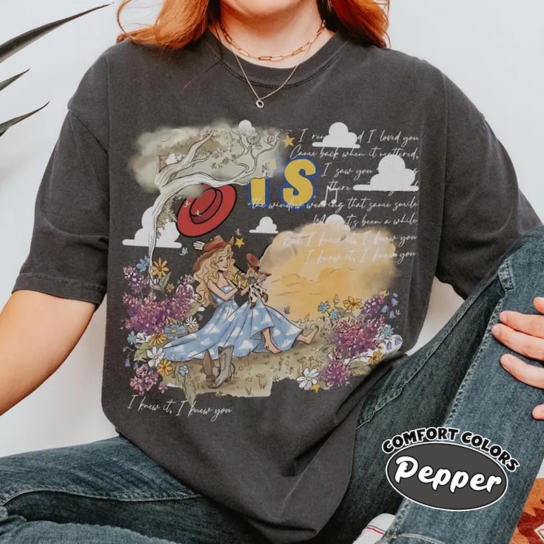 May include: A charcoal gray short-sleeve t-shirt featuring a graphic print of a person with long blonde hair sitting in a field of purple and yellow flowers. The design includes a red hat, clouds, a tree, and the text "I knew it, I knew you."