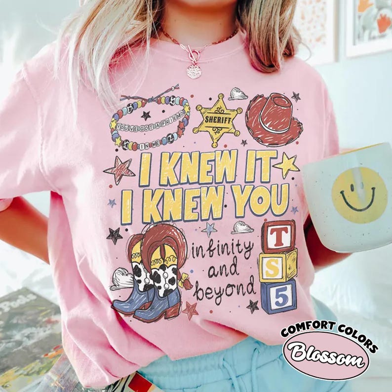 May include: A light pink t-shirt featuring a graphic print with the text I KNEW IT I KNEW YOU and infinity and beyond. The design includes a beaded bracelet, a sheriff star, a cowboy hat, cow print boots, and three stacked alphabet blocks.