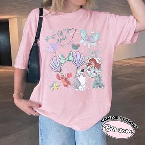 May include: A light pink t-shirt featuring a graphic print with the text Part Of Your World, a mermaid character, a crab, a fish, a dog, a seashell headband, a butterfly, and balloons. The design includes various pastel-colored illustrations.