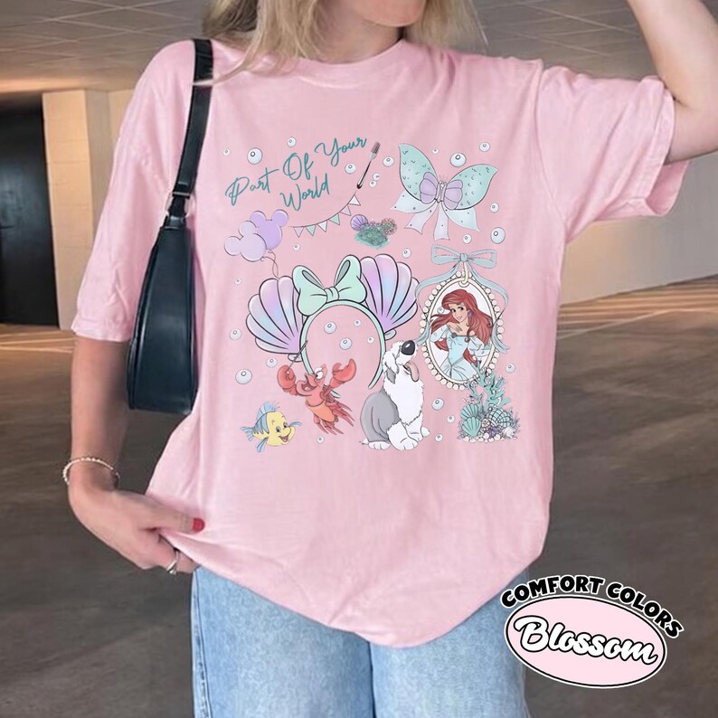 May include: A light pink t-shirt featuring a graphic print with the text Part Of Your World, a mermaid character, a crab, a fish, a dog, a seashell headband, a butterfly, and balloons. The design includes various pastel-colored illustrations.