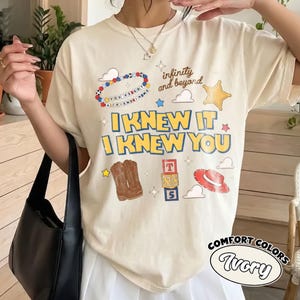 Pode incluir: Uma t-shirt cor de marfim com uma estampa gr&aacute;fica com o texto I KNEW IT I KNEW YOU, infinity and beyond, e YOU'VE GOT A FRIEND IN ME. O design inclui ilustra&ccedil;&otilde;es de uma pulseira de contas, uma estrela de xerife, botas, blocos e um chap&eacute;u.