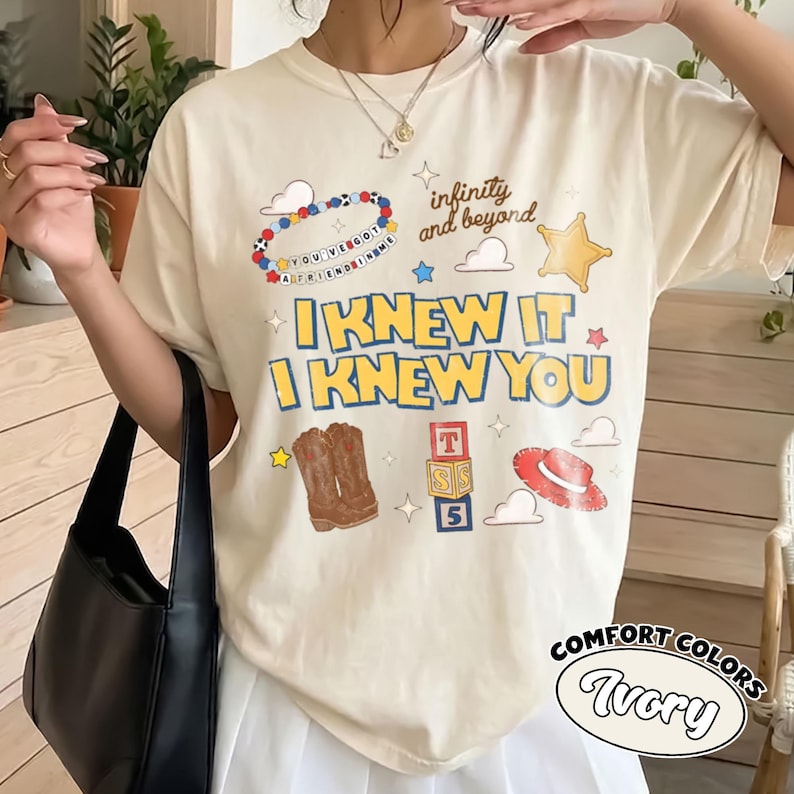 Pode incluir: Uma t-shirt cor de marfim com uma estampa gr&aacute;fica com o texto I KNEW IT I KNEW YOU, infinity and beyond, e YOU'VE GOT A FRIEND IN ME. O design inclui ilustra&ccedil;&otilde;es de uma pulseira de contas, uma estrela de xerife, botas, blocos e um chap&eacute;u.