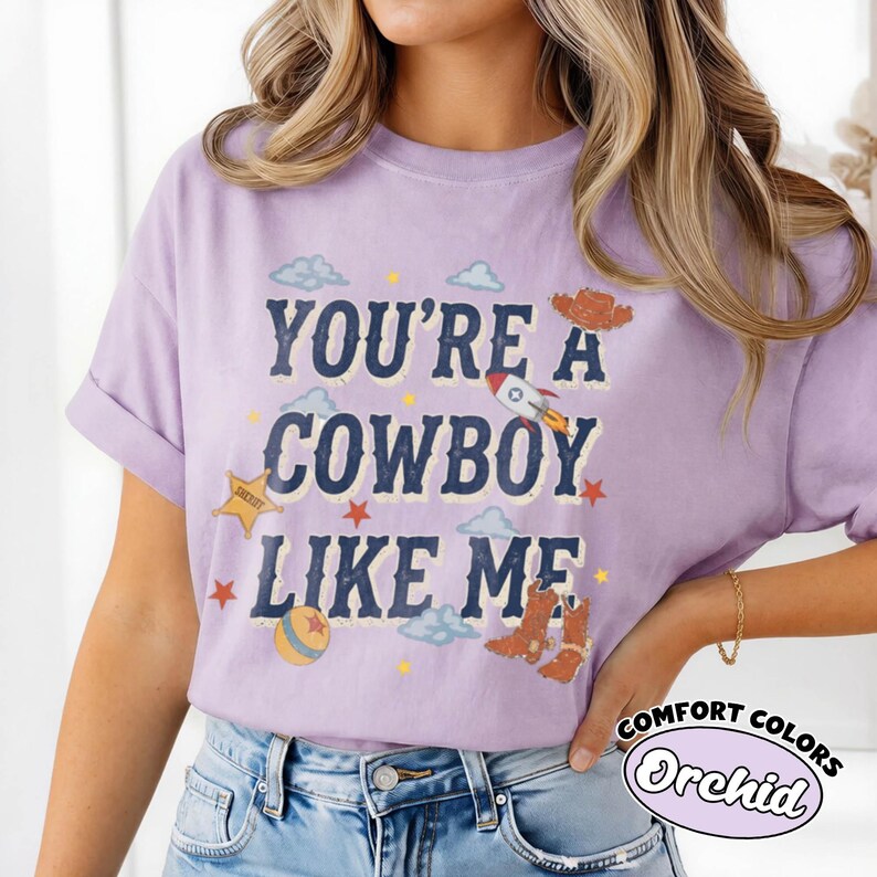 May include: A light purple short-sleeve t-shirt featuring the dark blue text "YOU'RE A COWBOY LIKE ME" surrounded by graphic illustrations of a rocket, a sheriff star, a ball, cowboy boots, a cowboy hat, and small blue clouds and stars.